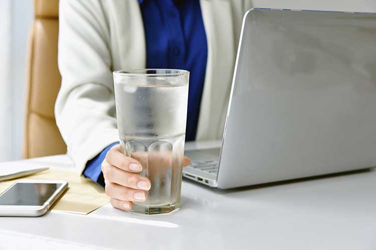 woman holding a glass of water on a desk to drink to prevent uti while working on a computer
